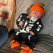Zev joined the competition — help win amazing prizes! baby, infant, child, portrait, sitting, pacifier, skeleton_costume, pumpkin, halloween, orange_hat, fuzzy_slippers, onesie, quilted_blanket, cushion, indoors, sleeping_area, hand, face, cute, seasonal