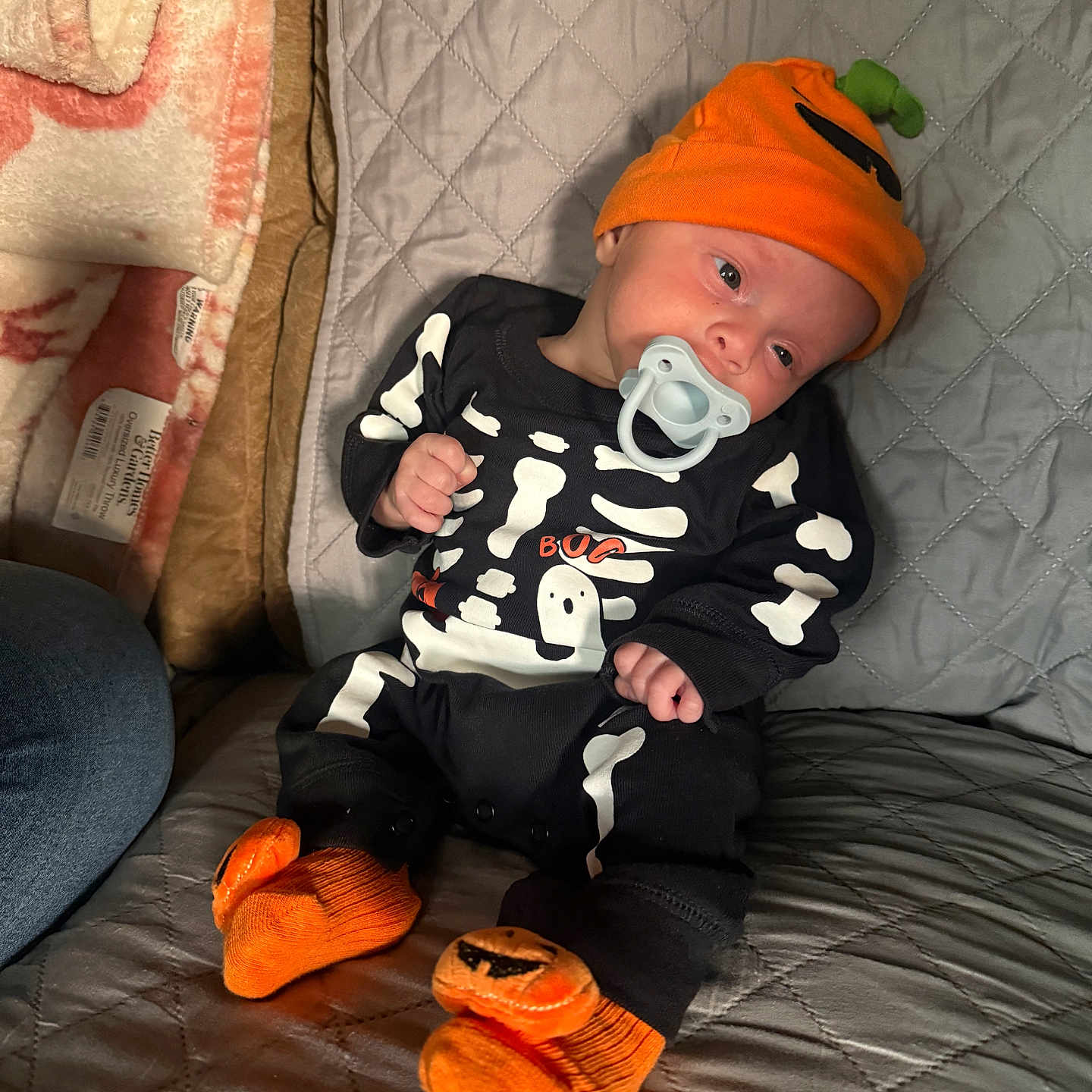 Zev joined the competition — help win amazing prizes! baby, infant, child, portrait, sitting, pacifier, skeleton_costume, pumpkin, halloween, orange_hat, fuzzy_slippers, onesie, quilted_blanket, cushion, indoors, sleeping_area, hand, face, cute, seasonal