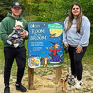 Hudson is registered to the contest to win money with this photo: man, baby, woman, dog, outdoor, forest, trail_sign, greenery, casual_clothing, hat, sneakers, leash, nature, family, walking, path, smile, holding, trees, daytime