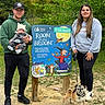 man, baby, woman, dog, outdoor, forest, trail_sign, greenery, casual_clothing, hat, sneakers, leash, nature, family, walking, path, smile, holding, trees, daytime