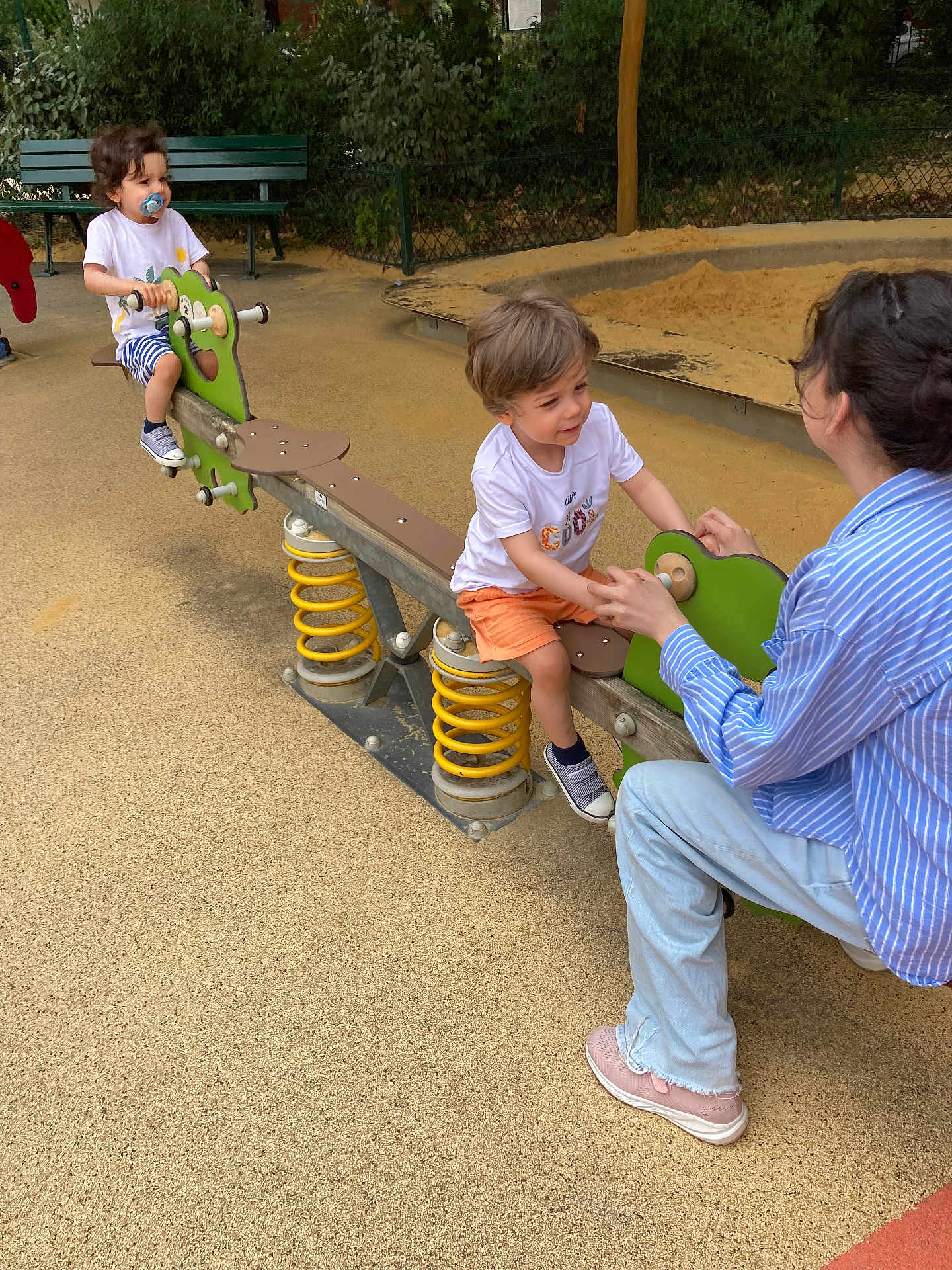 Axel Et Leo a rejoint le concours — aidez-le/la à gagner de superbes lots ! adult, bench, casual_clothing, child, children, footwear, greenery, happy, interaction, outdoor, pacifier, playground, playing, sandpit, seesaw, shorts, smiling, spring, toy, tshirt
