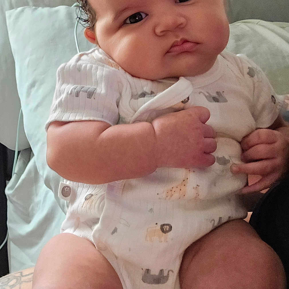Makaia is registered to the contest to win money with this photo: adult, baby, bedding, chubby_cheeks, closeup, curly_hair, cute, expression, eyes, face, hands, indoor, infant, legs, onesie, pillow, portrait, sitting, skin, tattoo