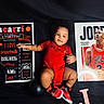 Jacarri joined the competition — help win amazing prizes! baby, basketball, black_background, black_hair, child, cute, indoors, infant, laying_down, letter_j, michael_jordan, milestone_board, photography, playful, portrait, poster, red_clothing, smiling, sneakers, sports_theme