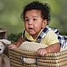 baby, child, basket, curly_hair, yellow_shirt, checkered_overalls, plush_toy, green_background, indoor, cute, infant, hands, face, smiling, soft_lighting, toy, floor, woven, curious, portrait