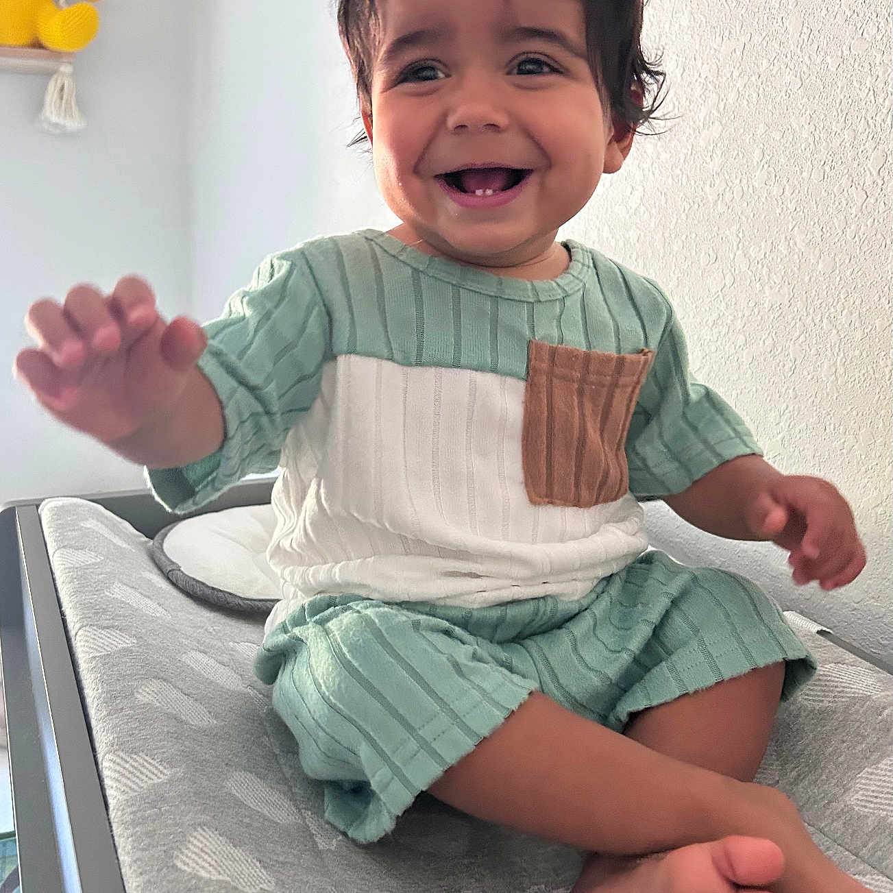 Adriel is registered to the contest to win money with this photo: toddler, child, smiling, happy, sitting, indoor, clothing, green_outfit, changing_table, baby_teeth, hand, foot, hair, face, toy, stuffed_animal, bear, shelf, wall, cushion