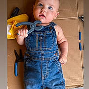 Roman is registered to the contest to win money with this photo: baby, child, denim_overalls, tool, clamp, cardboard, yellow_cloth, pliers, floor, curious, blue_eyes, expression, hand, foot, casual, indoor, portrait, infant, cute, playful