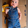 baby, child, denim_overalls, tool, clamp, cardboard, yellow_cloth, pliers, floor, curious, blue_eyes, expression, hand, foot, casual, indoor, portrait, infant, cute, playful