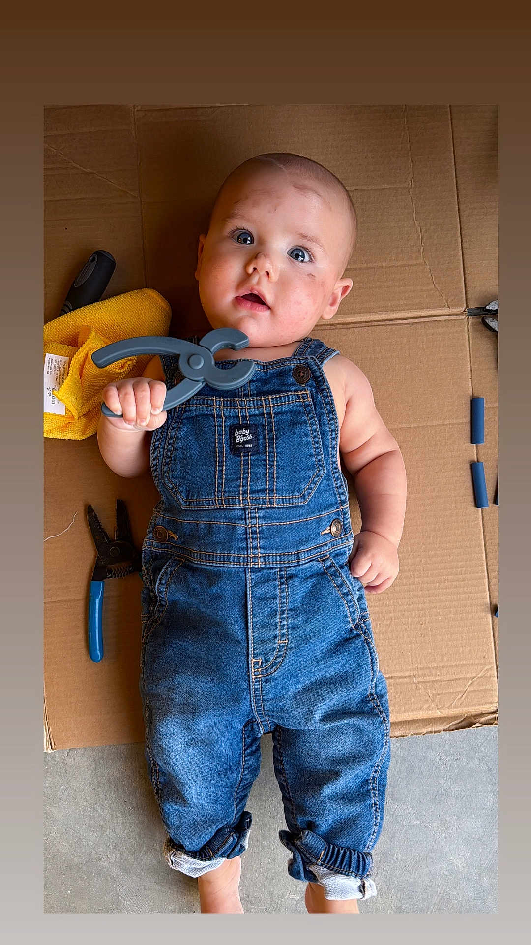 Roman is registered to the contest to win money with this photo: baby, child, denim_overalls, tool, clamp, cardboard, yellow_cloth, pliers, floor, curious, blue_eyes, expression, hand, foot, casual, indoor, portrait, infant, cute, playful