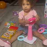 Ziah Rush is registered to the contest to win money with this photo: carpet, casual_clothing, child, chips, couch, curly_hair, floor, indoor, living_room, pink, playtime, shoes, shorts, sitting, snack, stacking_toy, table, toddler, toy, wooden_floor
