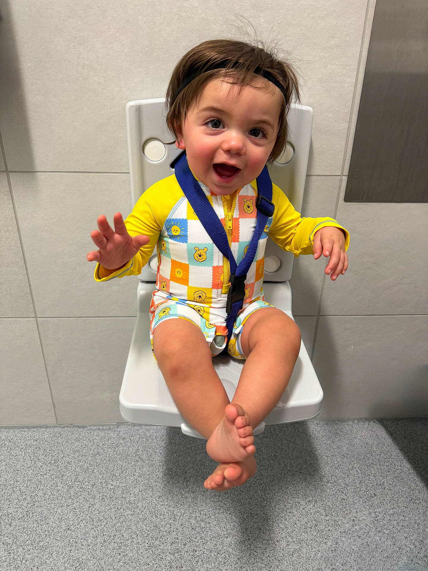 baby, barefoot, chair, child, colorful_clothing, cute, face, floor, happy, headband, indoor, onesie, person, playful, seat, smiling, strapped, toddler, wall, waving_hands
