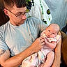 Adaline joined the competition — help win amazing prizes! man, baby, glasses, blanket, indoor, chair, tattoo, person, holding, infant, pink_clothing, facial_expression, curly_hair, comfort, parent_child, casual_clothing, portrait, looking_down, seated, family