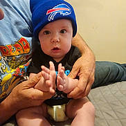 Calvin is registered to the contest to win money with this photo: adult_hands, baby, bed, bedroom, black_shirt, blue_beanie, casual, child, clothing, curious_expression, cute, diaper, headwear, holding, indoor, infant, person, sitting, skin, tshirt