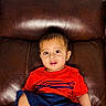 toddler, child, red_shirt, blue_shorts, brown_couch, indoor, sitting, curious, wide_eyes, leather, foot, hand, portrait, casual, young_child, home, furniture, cute, skin, person
