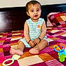 baby, bed, bedspread, child, colorful, cute, face, footwearless, head, indoor, infant, person, playful, quilt, sitting, smiling, striped_clothing, toy, wooden_headboard, young_child