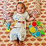baby, child, infant, toys, bedspread, pattern, clothing, lion_king, smiling, cute, lying_down, colorful, playful, indoors, face, hands, feet, pillow, fabric, expression