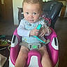 baby_seat, carpet, child, curly_hair, expression, furniture, green, holding, indoor, infant, kitchen, living_room, person, pink, seat_belt, toddler, toy, white, window, wooden_cabinet