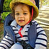baby, blue_pants, casual_clothing, child, daylight, face, happy, hat, nature, outdoor, portrait, red_strap, seatbelt, smiling, striped_sweater, stroller, sunlight, toddler, trees, young_child