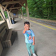 Younis participe au concours pour gagner de l'argent avec cette photo : child, boy, thumbs_up, train_station, platform, wooden_bench, trees, greenery, casual_clothing, tshirt, sneakers, outdoor, daylight, smile, hair, concrete_floor, wooden_structure, railway_tracks, nature, portrait