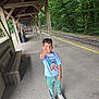 child, boy, thumbs_up, train_station, platform, wooden_bench, trees, greenery, casual_clothing, tshirt, sneakers, outdoor, daylight, smile, hair, concrete_floor, wooden_structure, railway_tracks, nature, portrait
