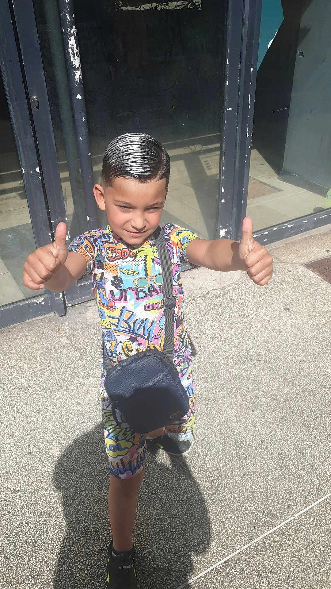Younis a rejoint le concours — aidez-le/la à gagner de superbes lots ! child, boy, thumbs_up, colorful_clothing, shorts, sneakers, bag, hair, smile, outdoor, sunlight, shadow, concrete, glass_door, urban, standing, happy, casual, person, daylight
