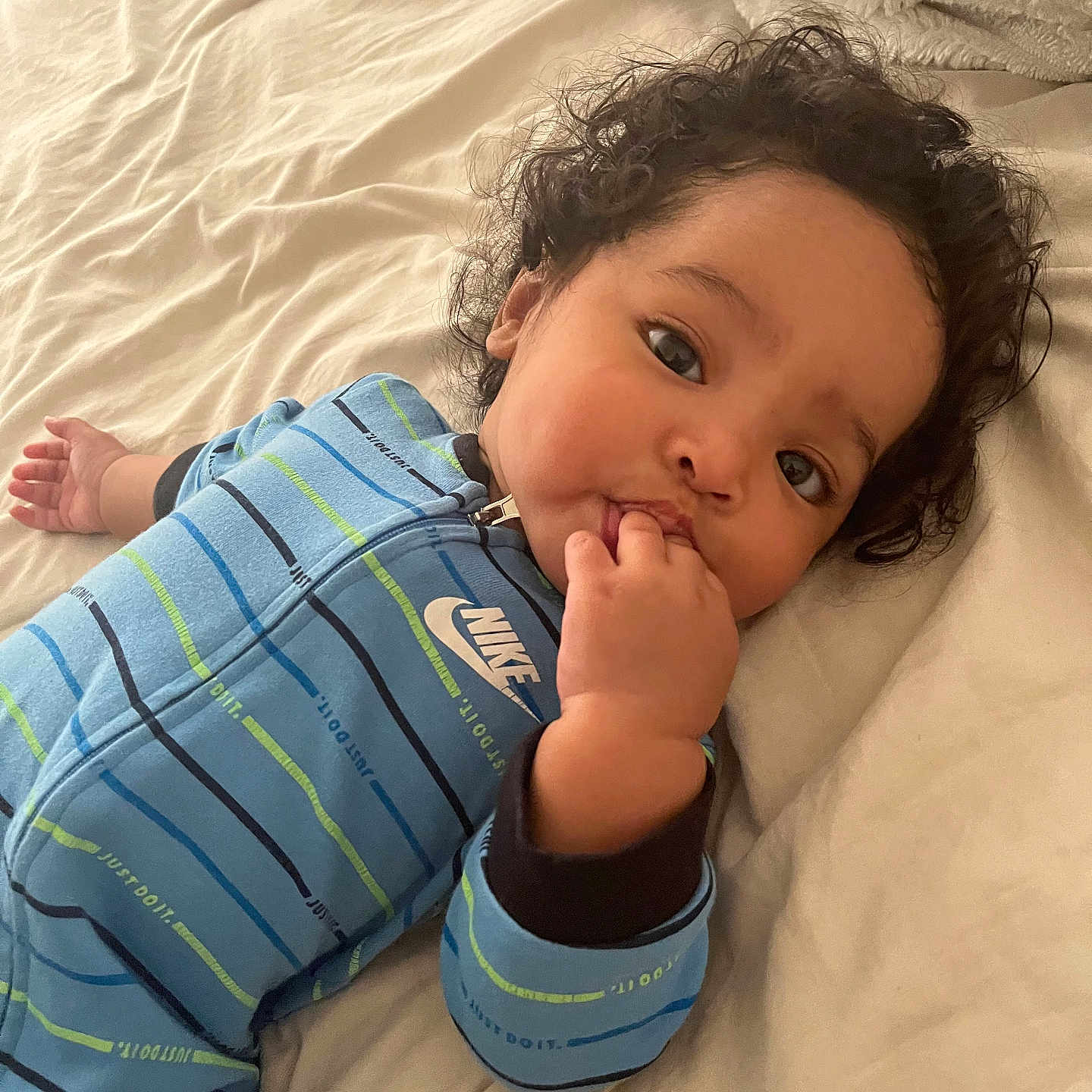 Cainnan is registered to the contest to win money with this photo: baby, bed, bedding, blue_onesie, child, cozy, curly_hair, cute, fingers_in_mouth, indoors, infant, lying_down, nike, person, portrait, sleepwear, sleepy, soft_texture, striped_clothing, young_child
