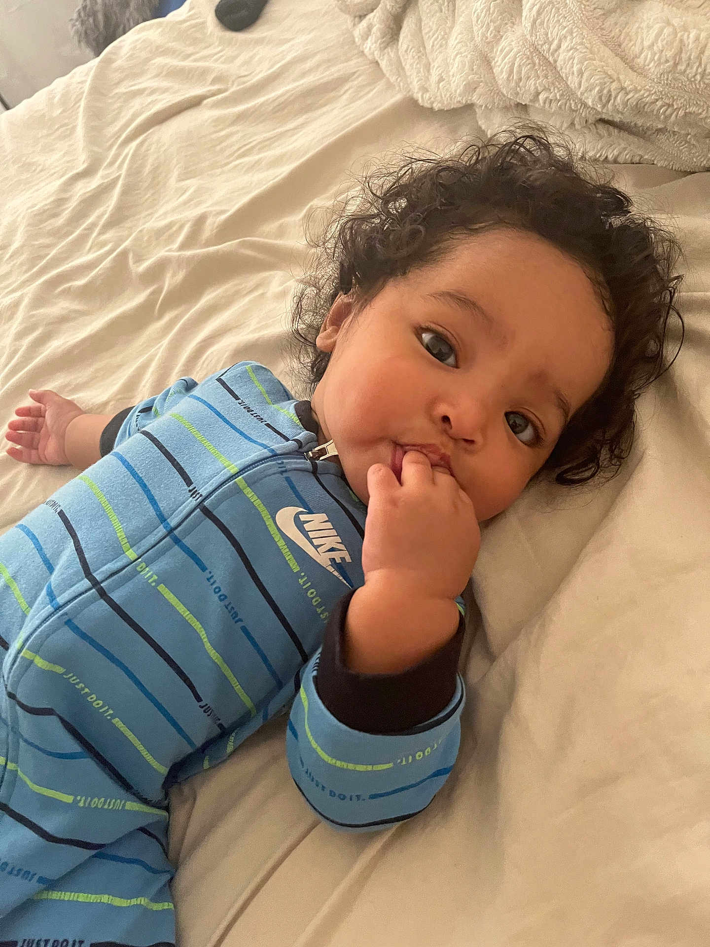 Cainnan is registered to the contest to win money with this photo: baby, child, curly_hair, blue_onesie, nike, bed, bedding, sleepwear, infant, lying_down, cute, fingers_in_mouth, soft_texture, striped_clothing, person, indoors, portrait, sleepy, young_child, cozy