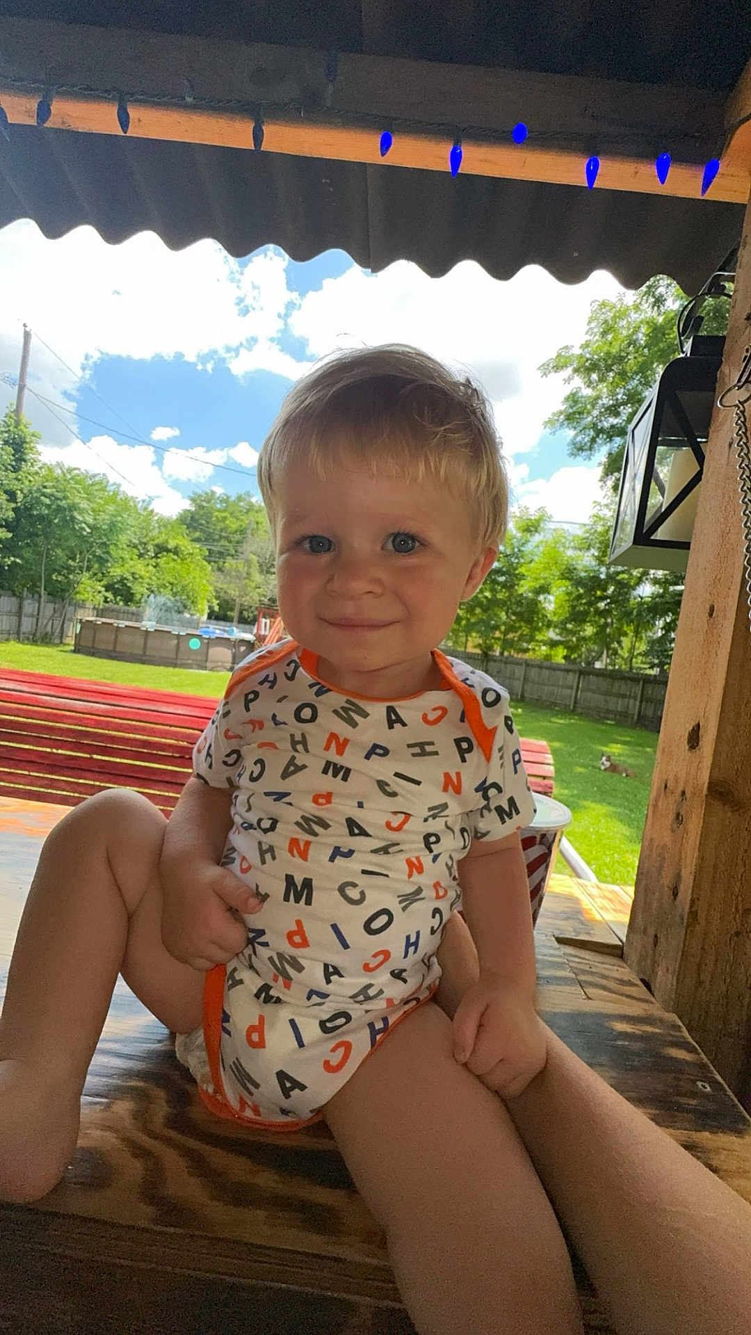 Dakota Jr is registered to the contest to win money with this photo: toddler, child, smiling, onesie, alphabet_pattern, bench, wood, outdoor, backyard, grass, trees, fence, pool, sky, clouds, sunny, face, person, daylight, shade