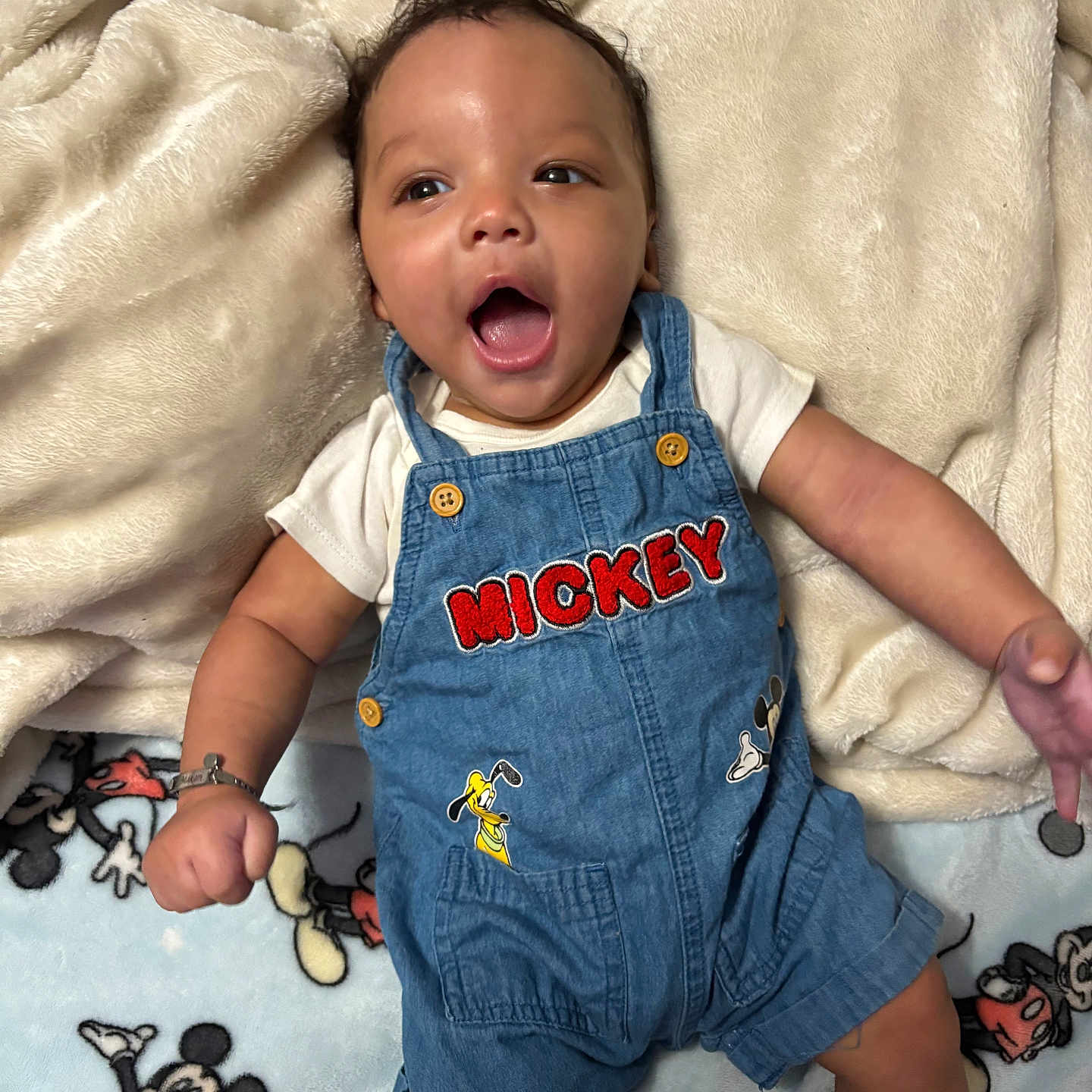 Makari is registered to the contest to win money with this photo: baby, child, denim_overalls, mickey_mouse, blanket, smiling, happy, infant, cute, clothing, portrait, lying_down, soft_texture, fabric, person, button, toy_character, arm, face, skin