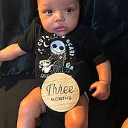 Makari joined the competition — help win amazing prizes! baby, infant, child, onesie, black_clothing, wooden_sign, text, three_months, sitting, portrait, indoor, cute, face, hands, legs, skin, expression, dark_background, photography, person