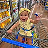 baby, child, shopping_cart, grocery_store, juice, yellow_outfit, headband, smiling, happy, waving, aisle, floor, shoes, person, retail, shopping, supermarket, cart_handle, sitting, infant