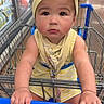 baby, child, shopping_cart, yellow_headband, striped_outfit, supermarket, aisle, floor, hand, face, expression, clothing, footwear, indoor, person, retail, grocery_store, cart_handle, cute, toddler