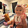 baby, child, smiling, headband, bib, stuffed_animal, bed, striped_bedspread, toy, indoor, room, furniture, dresser, curtains, hat, socks, person, portrait, cute, happy