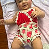 baby, infant, smile, happy, towel, clothing, romper, watermelon_print, floral_trim, curly_hair, skin, face, arms, legs, wooden_floor, indoors, cute, lying_down, child, person