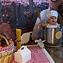 baby, child, chef_hat, cooking_pot, wooden_spoon, spaghetti, bread, basket, checkered_cloth, toy_mouse, kitchen_theme, table, floor, decor, indoor, playful, smiling, portrait, costume, background