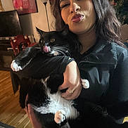 Francis joined the competition — help win amazing prizes! person, cat, indoor, black_cat, white_paws, wooden_floor, holding, licking_nose, puckered_lips, smiling, long_hair, ring, jacket, lamp, painting, television, chair, plant, home, portrait