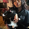 person, cat, indoor, black_cat, white_paws, wooden_floor, holding, licking_nose, puckered_lips, smiling, long_hair, ring, jacket, lamp, painting, television, chair, plant, home, portrait