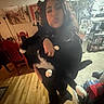 Francis is registered to the contest to win money with this photo: person, cat, indoor, kitchen, refrigerator, wooden_floor, red_chair, holding, black_cat, white_paws, magnet, note, furniture, appliance, home, casual, blurry, portrait, floor_tile, lamp