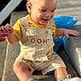 baby, child, concrete, curly_hair, earring, footwear, hand_holding, happy, laughing, outdoor, overalls, person, pooh, sitting, smile, sneakers, socks, sunlight, toddler, yellow_shirt