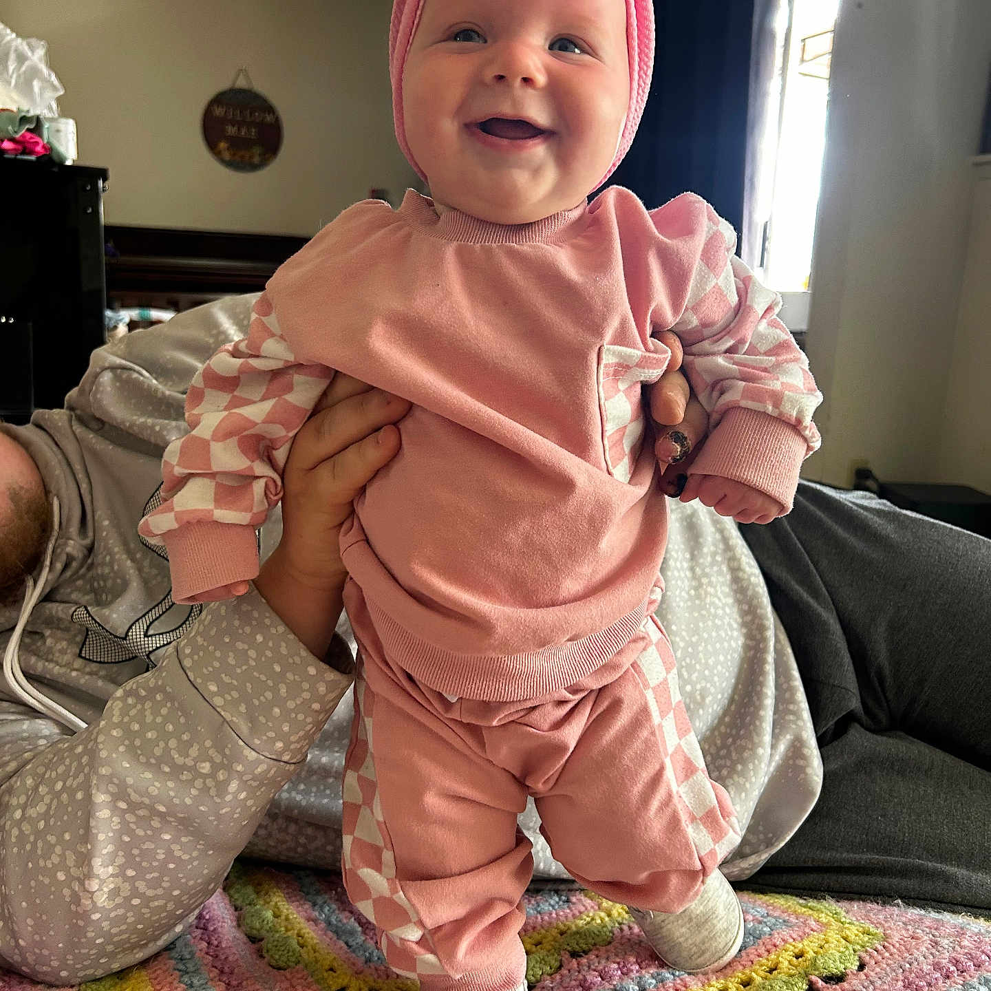 Willow Mae is registered to the contest to win money with this photo: adult_hand, baby, blanket, casual_wear, cheerful, child, cozy, crocheted, face, footwear, happy, headband, holding, home, human, indoor, infant, person, pink_clothing, smile