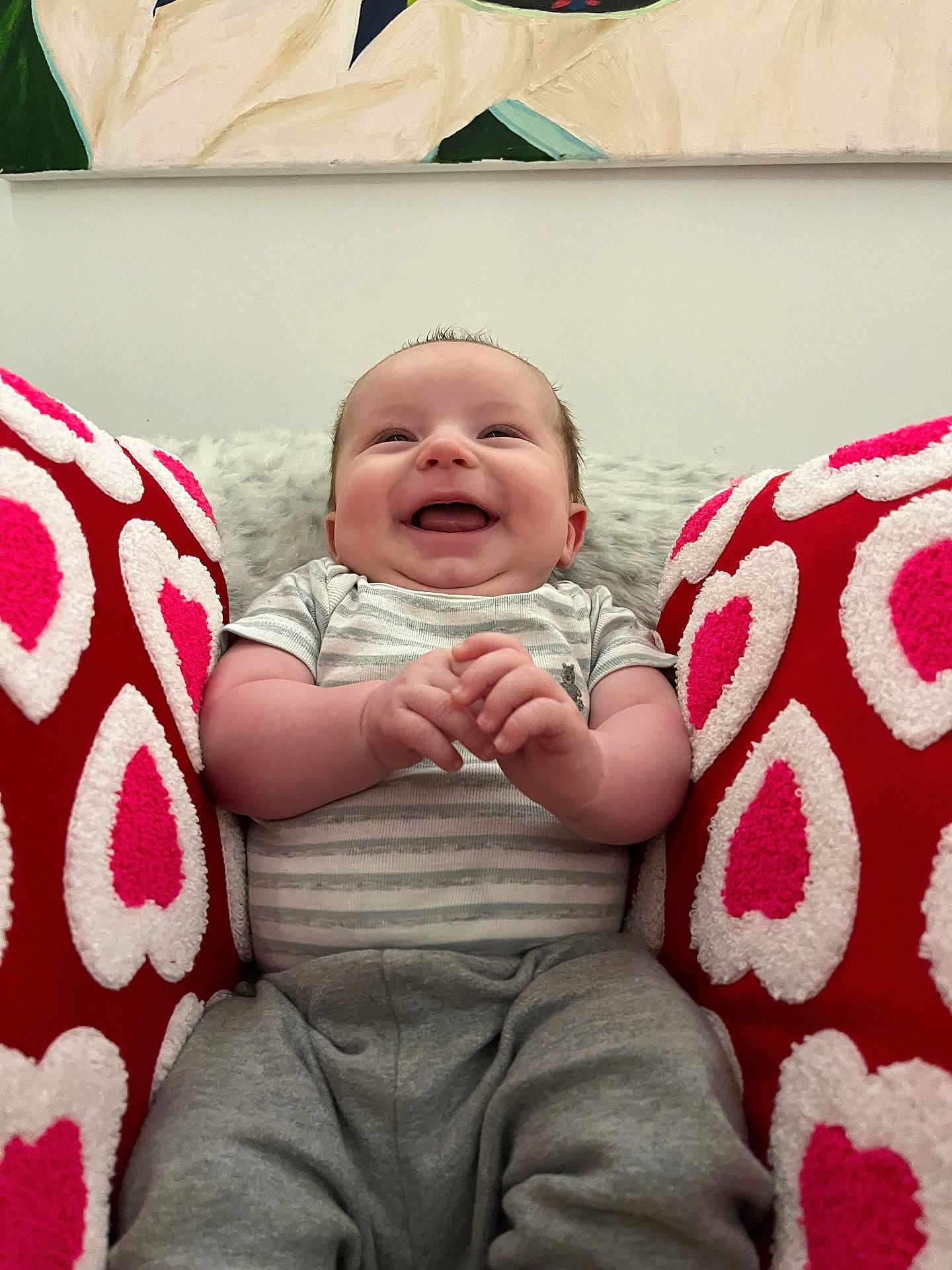 Onyx is registered to the contest to win money with this photo: baby, infant, smiling, laughing, face, hands, onesie, striped_onesie, gray_pants, red_pillows, heart_pattern, cushion, plush_blanket, wall_art, indoor, portrait, seated, cozy, happy, chubby_cheeks