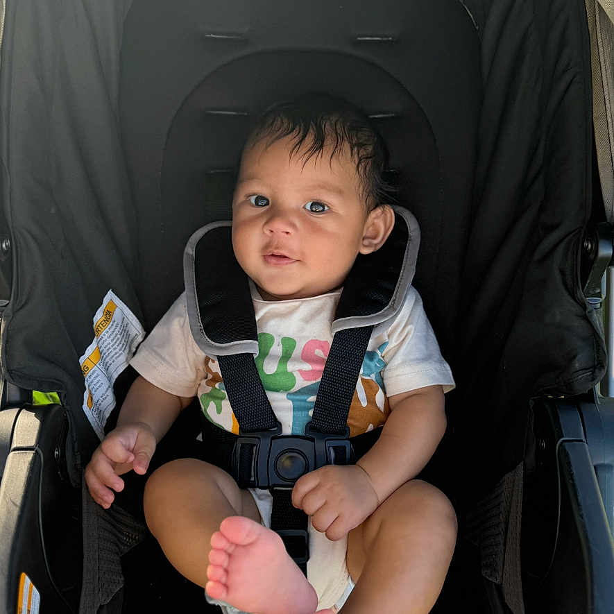 Lorenzo is registered to the contest to win money with this photo: baby, black_stroller, child, clothing, cute, face, foot, happy, head, infant, outdoor, person, portrait, safety_harness, seatbelt, sitting, smiling, stroller, sunlight, young
