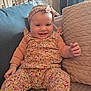 baby, smiling, child, floral_outfit, headband, couch, pillow, indoor, home, happy, person, sitting, cute, cozy, infant, patterned_clothing, living_room, portrait, soft_light, relaxed
