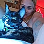 cat, tortoiseshell_cat, pet, person, selfie, indoor, couch, collar, bells, tattoo, smile, relaxing, lights, string_lights, bedroom, face, fur, whiskers, closeup, comfort