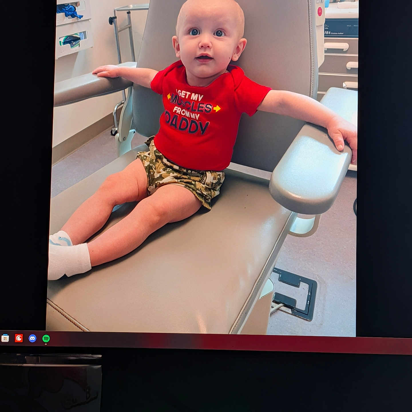 Melvin is registered to the contest to win money with this photo: baby, child, clinic, curious, cute, expression, furniture, healthcare, hospital_room, indoors, infant, medical_chair, medical_equipment, person, portrait, red_shirt, shorts, sitting, socks, young_child