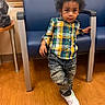 toddler, child, curly_hair, plaid_shirt, distressed_jeans, white_sneakers, wooden_floor, blue_chair, indoor, person, leaning, standing, casual_clothing, portrait, young_child, curious_expression, furniture, home_interior, flooring, waiting_room
