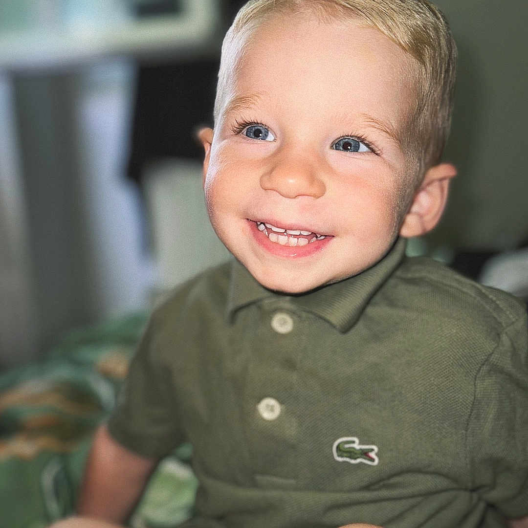 Nohé participe au concours pour gagner de l'argent avec cette photo : bed, blanket, blonde_hair, blue_eyes, casual, child, cute, face, green_shirt, happy, indoor, person, polo_shirt, portrait, shorts, sitting, skin, smiling, toddler, youth