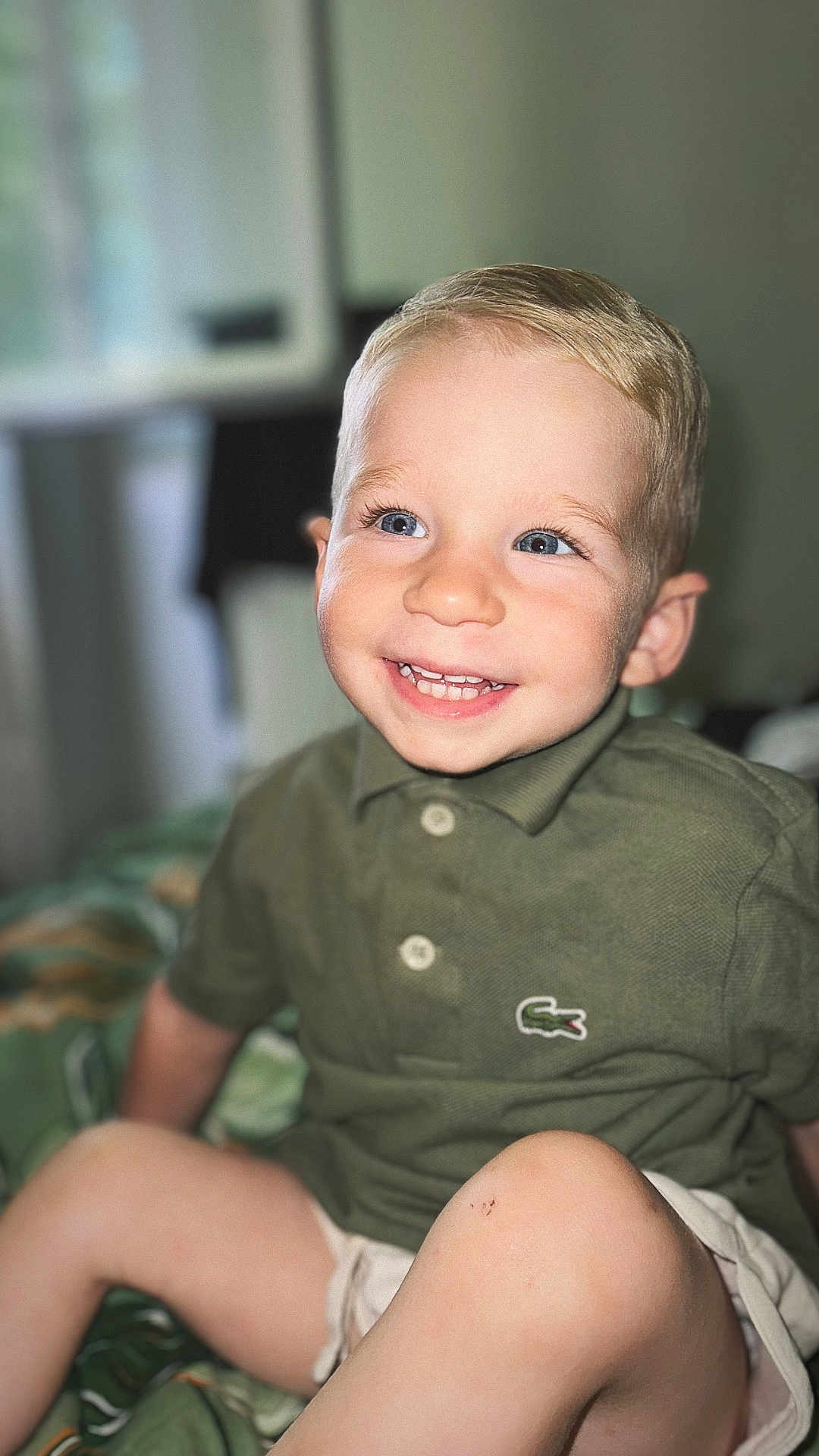 Nohé participe au concours pour gagner de l'argent avec cette photo : toddler, child, smiling, happy, blue_eyes, blonde_hair, polo_shirt, green_shirt, shorts, sitting, indoor, bed, blanket, face, person, cute, portrait, youth, skin, casual