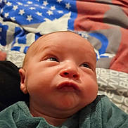 Malo participe au concours pour gagner de l'argent avec cette photo : baby, infant, newborn, face, eyes, nose, mouth, cheeks, hand, fingers, onesie, green_clothing, blanket, american_flag, bedding, bed, headboard, toy, portrait, indoor