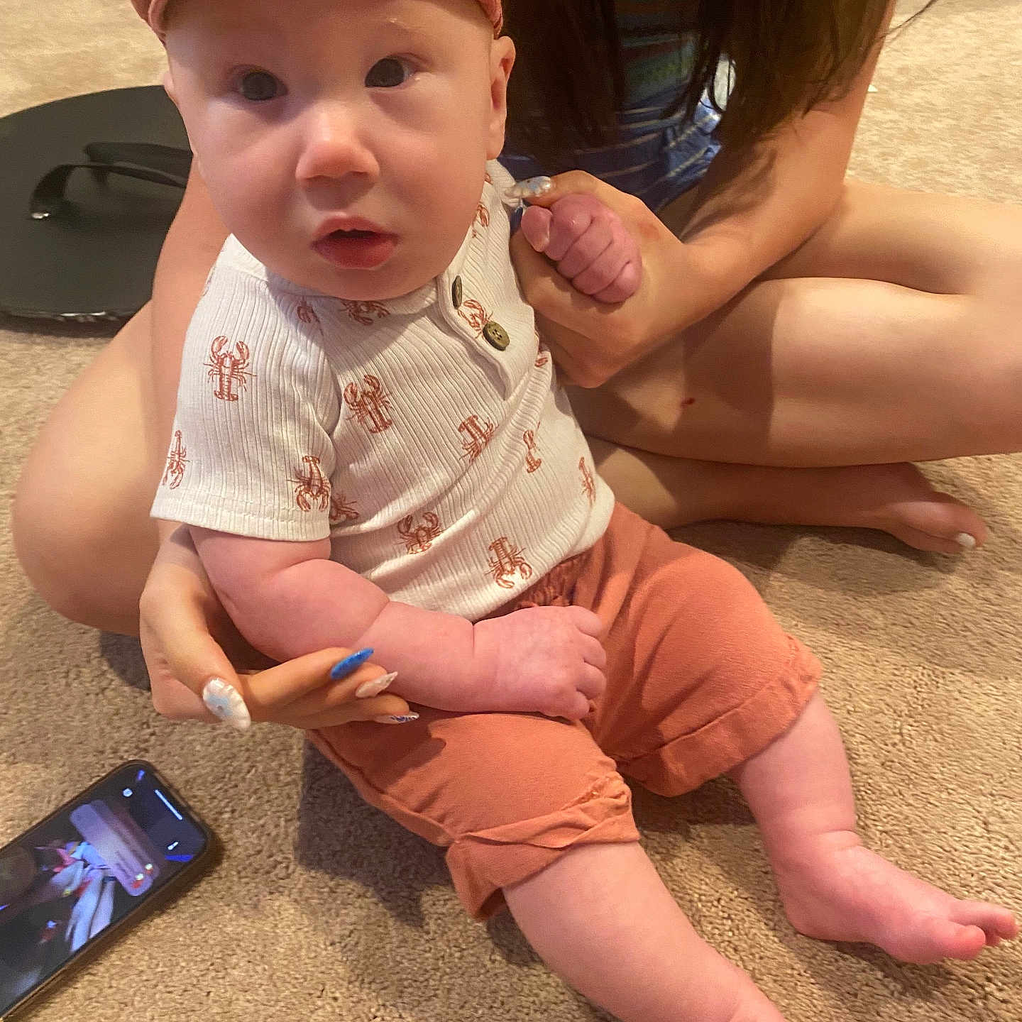 Grayson joined the competition — help win amazing prizes! adult, baby, cap, carpet, child, curious, face, feet, hand, holding, indoor, infant, legs, nails, pants, person, shirt, sitting, smartphone, video