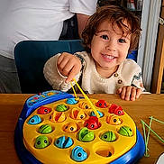 Aidan a rejoint le concours — aidez-le/la à gagner de superbes lots ! child, toddler, smiling, playing, toy, fishing_game, table, indoor, adult, fridge, white_shirt, wooden_table, happy, face, hand, colorful, game, furniture, person, hair